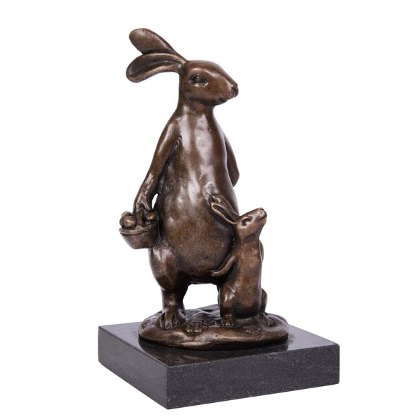 This is a bronze double-rabbit sculpture: the adult rabbit stands upright holding a basket, with a baby rabbit snuggling beside it, on a black stone base. It’s a warm family-themed decor piece.