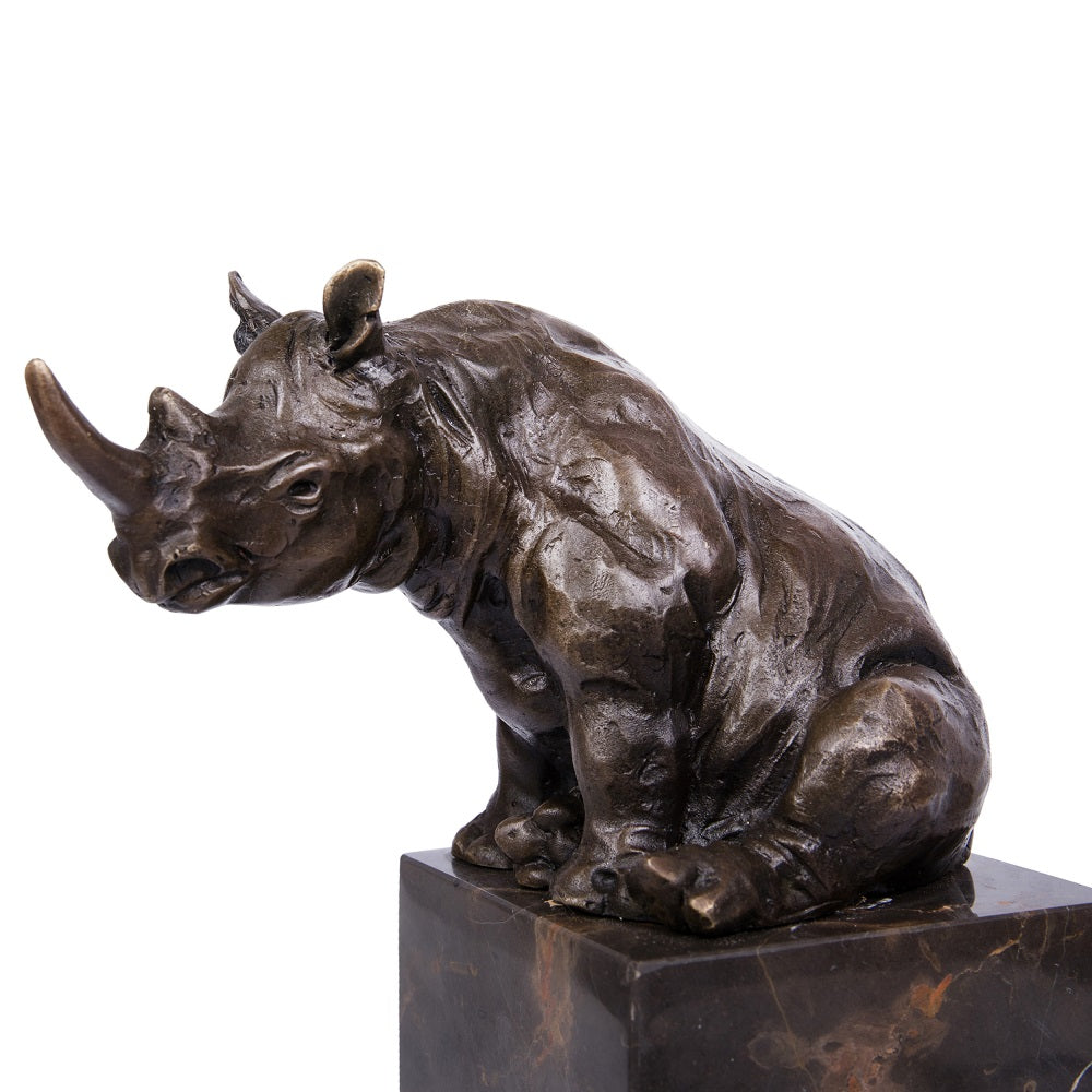 This is a bronze rhinoceros sculpture: It sits in a seated position, with a long horn extending forward and mouth slightly open. The rough, realistic texture of its body fully shows the rhino’s heavy quality. Placed on a textured black stone base, the sculpture conveys a calm sense of power overall.